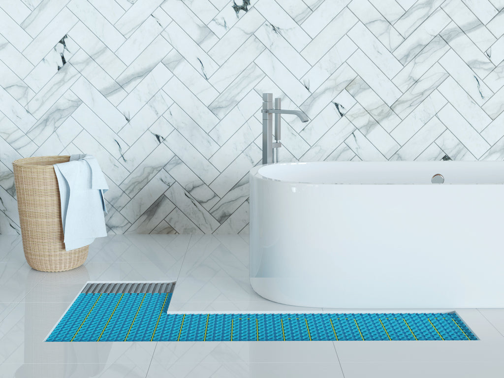 A blue and green striped L-shaped bath mat on a white tiled bathroom floor, positioned in front of a white freestanding bathtub and a gray floor drain, with a tile cutout showing the Thermosphere Ultimate underfloor heating in place.