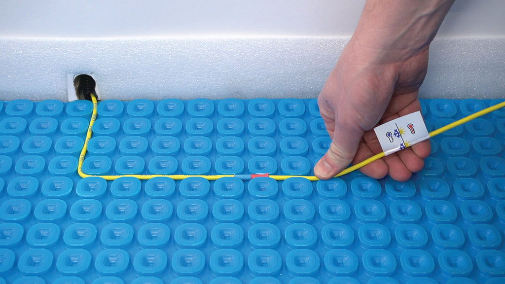 A close-up of a person's hand fixing a yellow heating wire into a blue Thermsophere ultimate mat with a small black component near the top left, against a white background.
