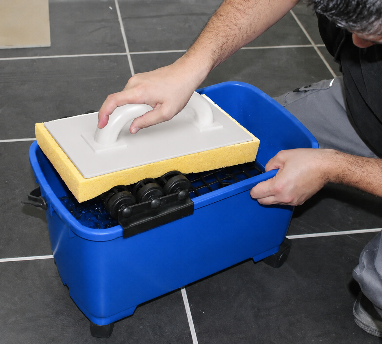 Tiler wringing MILLCOT grouting sponge float over blue wringer bucket system on dark slate tile floor