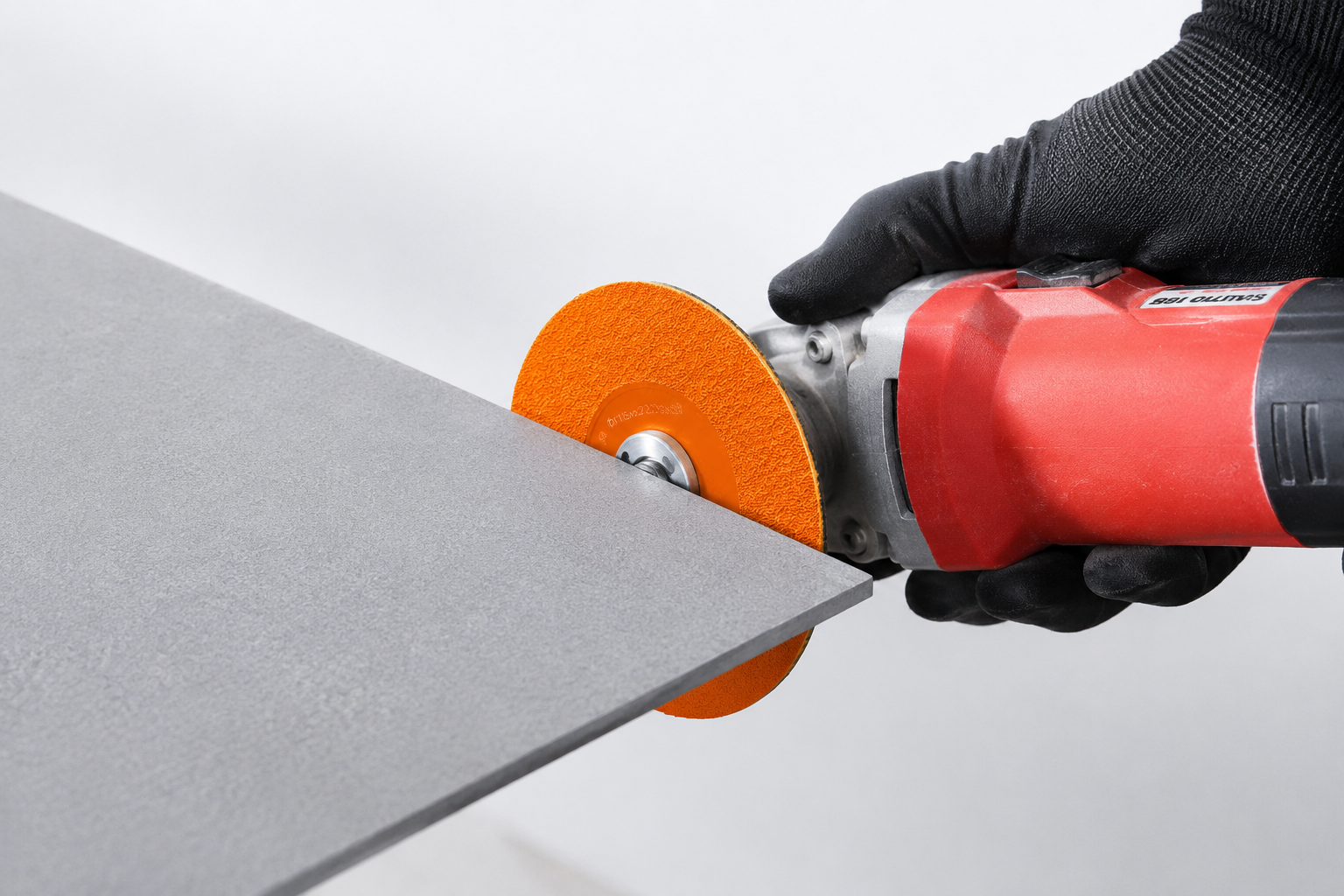 Close-up of gloved hands using a red angle grinder with the orange MILLCOT 115mm cutting and grinding disc to slice cleanly through the edge of a grey porcelain tile.
