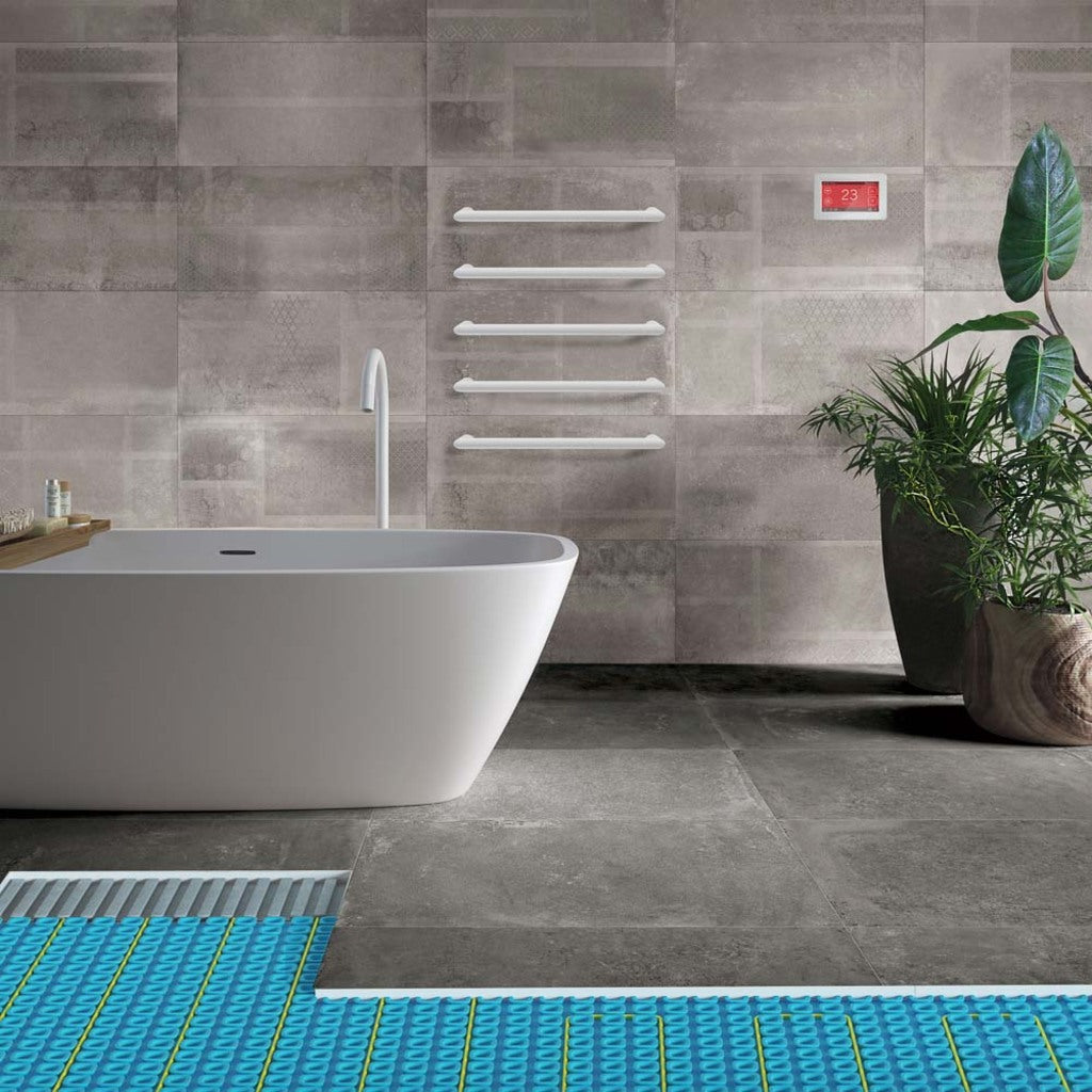 A modern bathroom scene with a freestanding white bathtub, gray tiled walls and floor, white towel bars, a small red digital thermostat on the wall, and a potted green plant. A blue patterned underfloor heating mat is visible in the foreground.