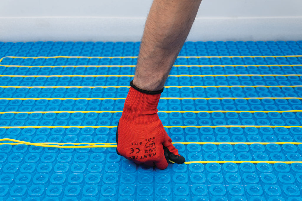 A close-up of a person's hand wearing a red work glove, pressing down on Thermosphere ultimate yellow heating cables to place it into the ultimate matting.