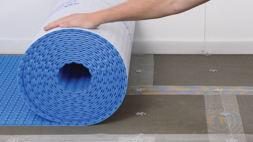 A person's arm is visible as they unroll a large roll of blue and white Thermosphere ultimate mat over a gray floor mesh on a white wall background.
