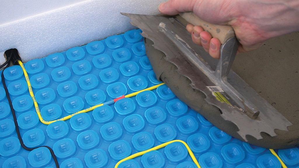A close-up of a person's hand using a trowel to apply tile adhesive over a blue Thermosphere ultimate mat with yellow and black heating cables.