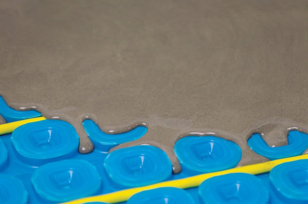 Close-up of a blue Thermosphere ultimate mat with a raised grid pattern, partially covered by wet gray self levelling compound, with yellow heating cables visible along the bottom.