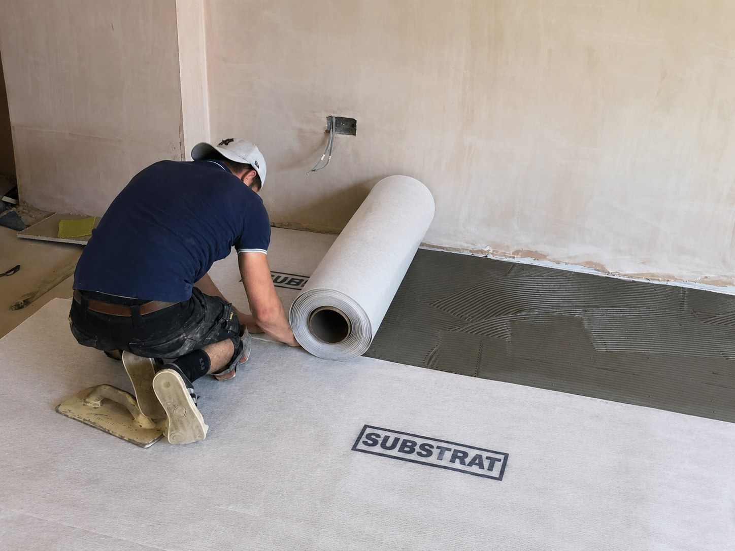 A worker kneels on a floor installation, unrolling a large roll of SUBSTRAT DecoupleX over a fresh bed of grey tile adhesive. The room has beige plaster walls.