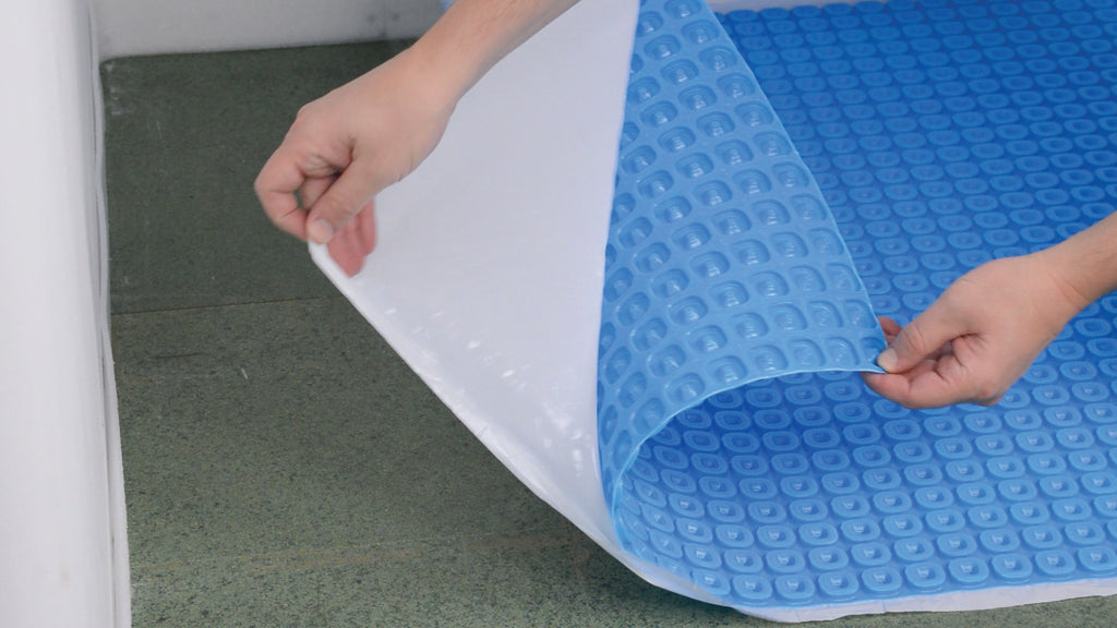 A person's hands peeling back a white protective film from a blue Thermosphere self adhesive ultimate matting for underfloor heating, ready to lay onto green chipboard flooring.